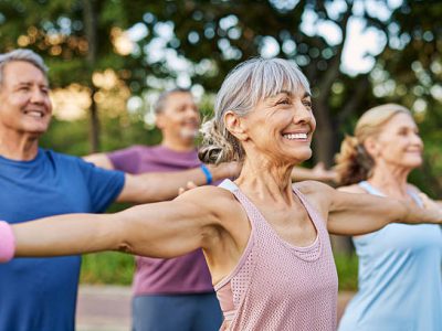 Happy senior group doing yoga together in the park, gentle gymnastics. Mature men and women stretching arms in outdoor fitness class. Sporty elderly friends doing a arm warm up workout before training class together in a garden.